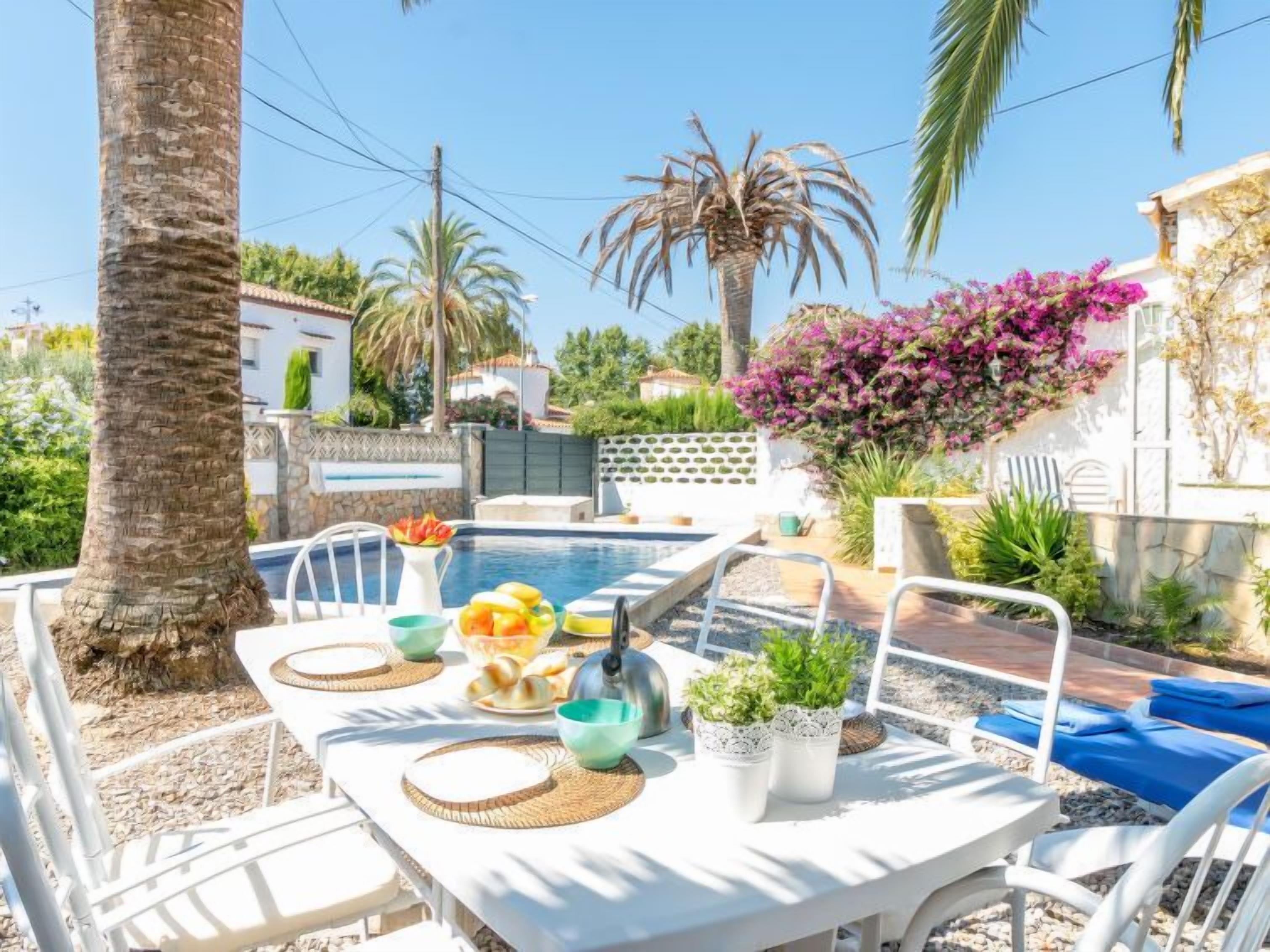 Un área de comedor junto a la piscina con una mesa preparada para dos, rodeada de palmeras y una piscina.