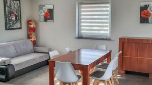 Una sala moderna con un sofá gris, una mesa de comedor de madera con cuatro sillas, y un mueble de madera. Hay dos cuadros enmarcados en las paredes y una ventana con persianas.