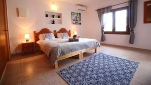 A bedroom with a wooden bed, bedside tables, a patterned rug, a window with curtains, and wall-mounted shelves with decorative items. - Villa Susan - Private & Peaceful | Pool, Wifi & AC (Sant Antoni de Portmany)