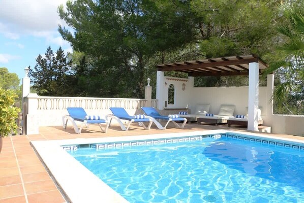 A swimming pool with blue water, surrounded by white walls and a pergola with a roof. There are lounge chairs with blue cushions and a white pillar with a decorative element.