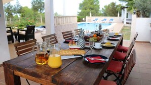 A wooden dining table set with plates, glasses, and a pitcher of juice, overlooking a swimming pool. - Villa Susan - Private & Peaceful | Pool, Wifi & AC (Sant Antoni de Portmany)