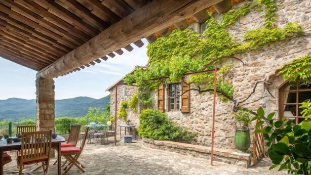 Un bâtiment en pierre doté d’une pergola en bois, d’un espace extérieur aménagé pour s’asseoir et offrant une vue sur les montagnes.