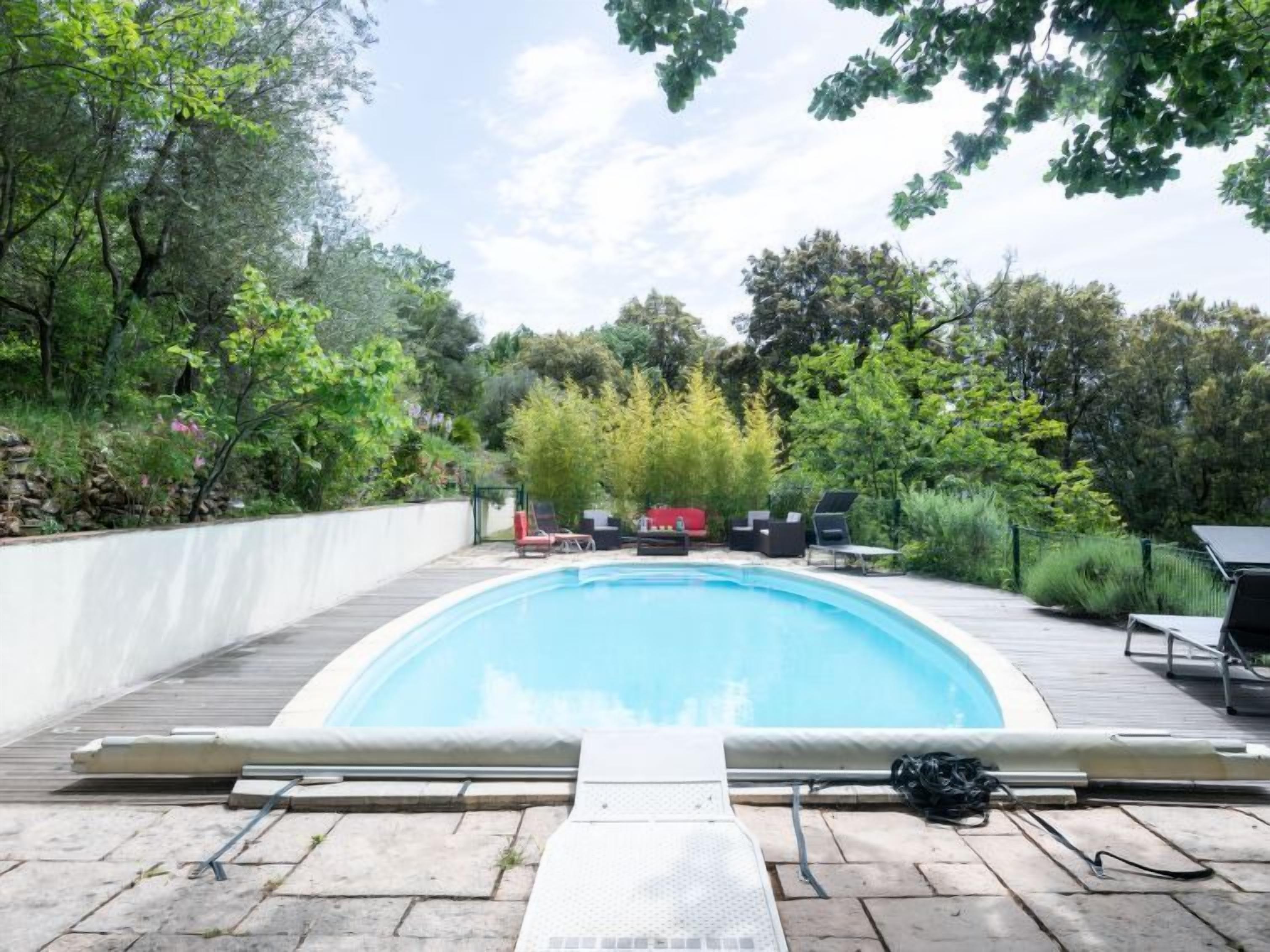 A rectangular swimming pool with a wooden deck, surrounded by lush greenery and trees.