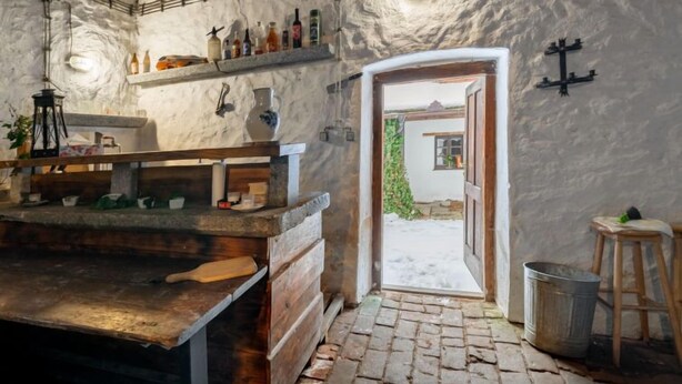 Eine rustikale Küche mit Steinwänden, einer Holzausstattung und einem Backsteinboden. An der Wand befindet sich ein Regal mit Flaschen und ein Laternen.