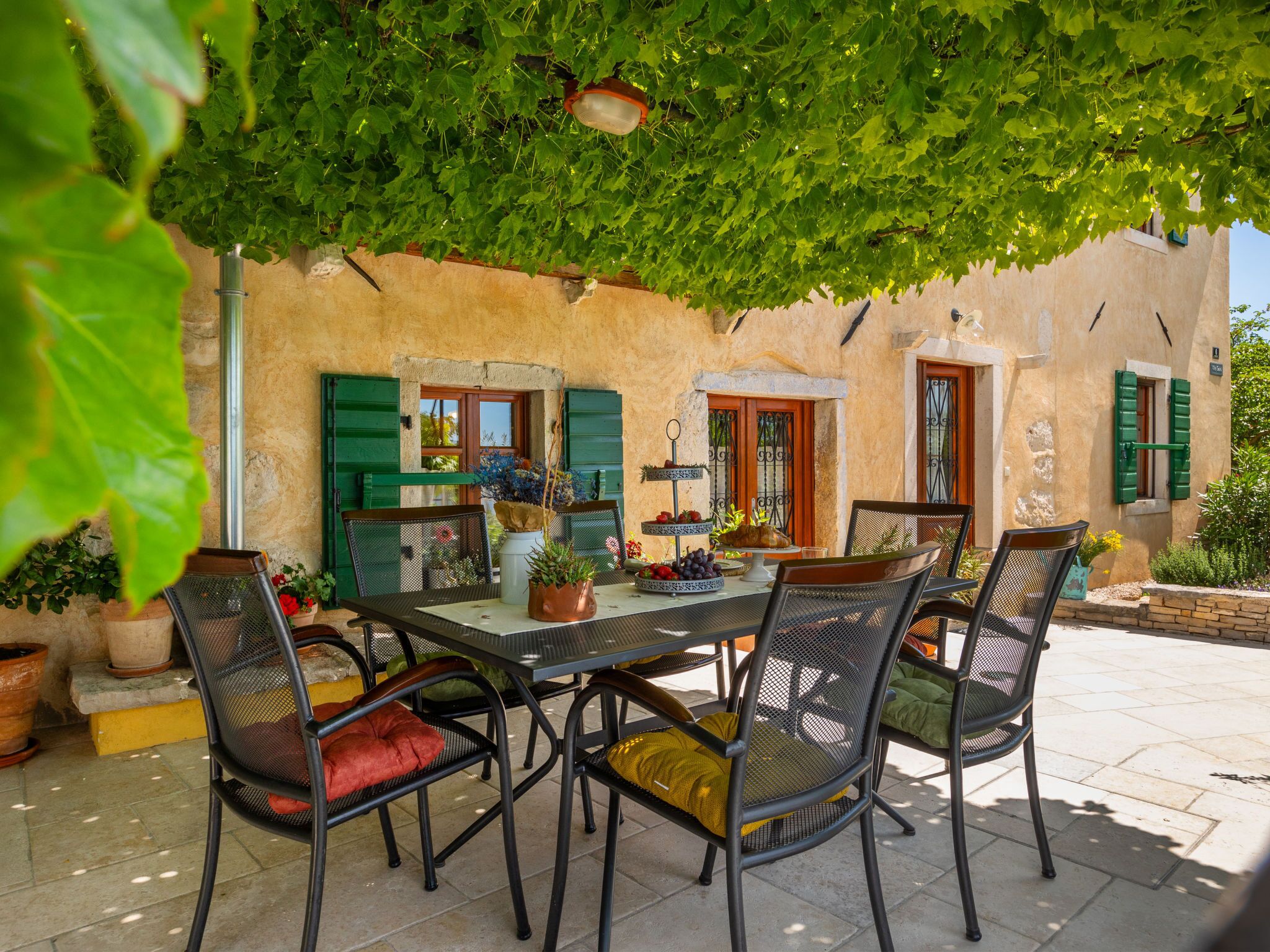 An outdoor dining area with a table and chairs, surrounded by potted plants and a building with green shutters.