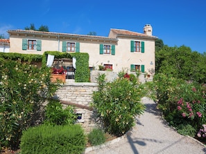 A two-story building with a stone pathway leading to the entrance, surrounded by lush greenery and flowering plants. - Vacation home Gelci by Interhome (Trget)