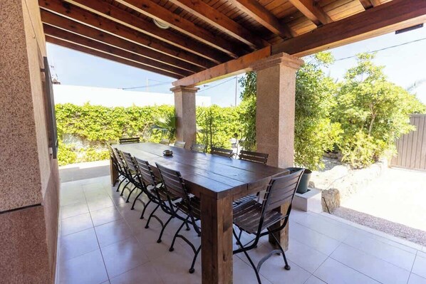An outdoor patio with a wooden table and chairs, a pergola overhead, and a view of greenery and a fence.
