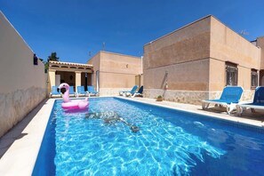A swimming pool with clear blue water, surrounded by lounge chairs and a building with a tiled roof.