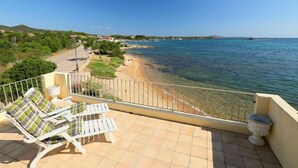 A balcony with two white lounge chairs and a potted plant, overlooking a beach with clear blue water and a distant shoreline. - Apartment Paola by Interhome (Cannigione)