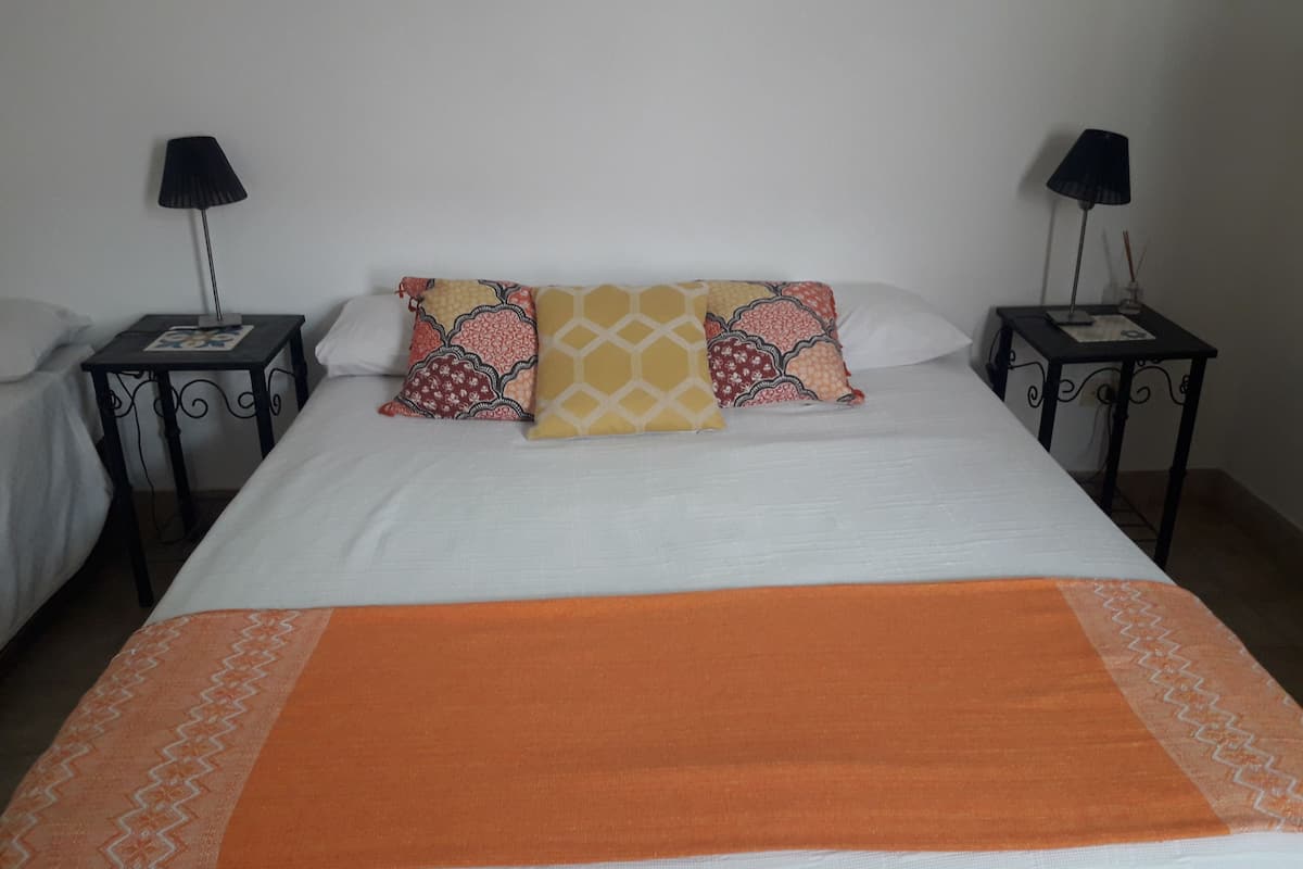 A neatly made bed with a white comforter, an orange throw blanket, and patterned pillows. There are two bedside tables with lamps and a small decorative item on each.