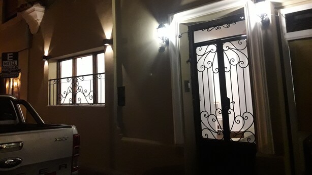 Entrada de hotel con una reja metálica decorativa, una camioneta pickup blanca estacionada afuera y un cartel que indica el nombre y los servicios del hotel.
