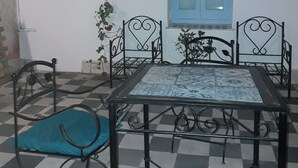 Una habitación con piso a cuadros, una mesa con mantel estampado, cuatro sillas, una ventana con contraventanas azules y una planta colgante.