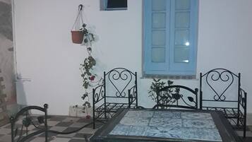 Una habitación con piso a cuadros, una mesa con mantel estampado, cuatro sillas, una ventana con contraventanas azules y una planta colgante.