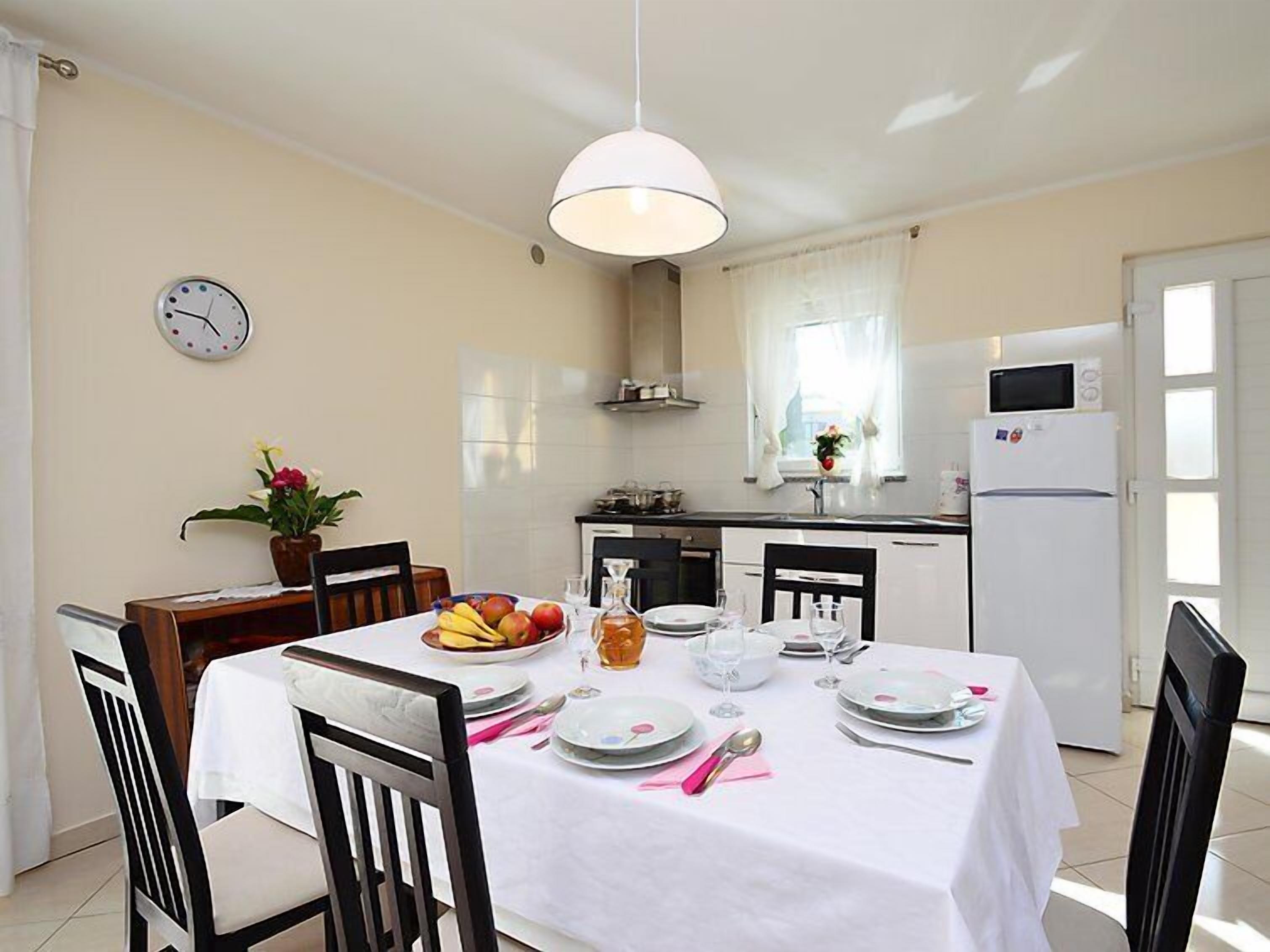 A dining area with a white table set for four, surrounded by chairs. A clock and a potted plant are on the wall.