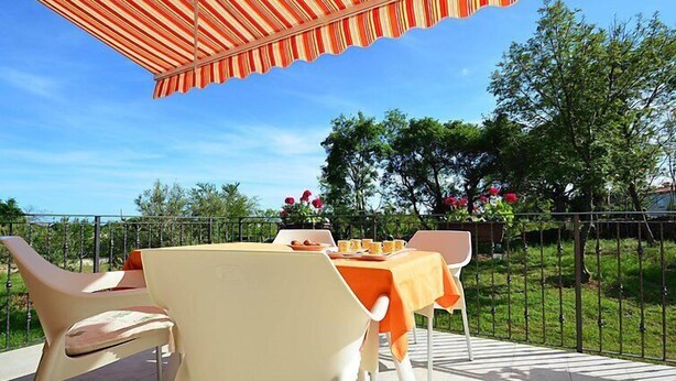 En udendørs terrasse med bord og stole, en stribet markise og udsigt til træer og et hegn.