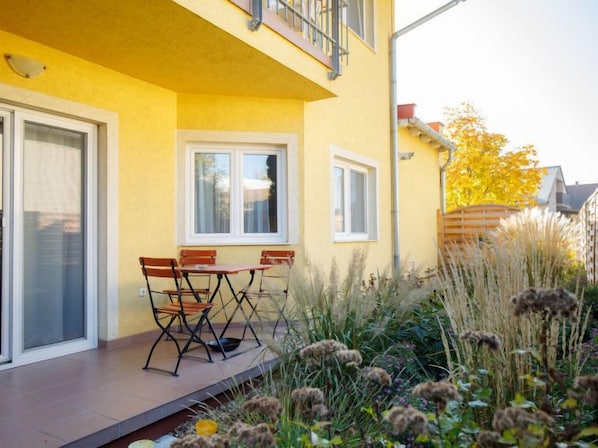 Un edificio amarillo con un balcón, una mesa con sillas y un jardín con pastos altos y flores.