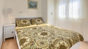 Une chambre à coucher avec une couvre-lit à motifs, une lampe de chevet, un tableau encadré au mur et une fenêtre ornée de rideaux légers.