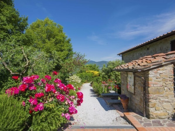 A stone building with a tiled roof, surrounded by lush greenery and a gravel path.