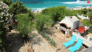 A patio with a stone pathway, a barbecue grill, and two blue lounge chairs overlooking a beach with waves and a flag.