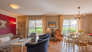 Un salon comprenant un canapé, des fauteuils, une table à manger avec des chaises, un lustre et une vue sur l’extérieur, visible à travers une porte vitrée.