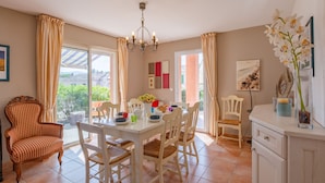 Un coin repas avec une table dressée pour quatre personnes, entourée de chaises blanches. On y trouve une chaise en bois avec un coussin à carreaux, un lustre au-dessus de la table et un vase rempli de fleurs sur le côté droit.