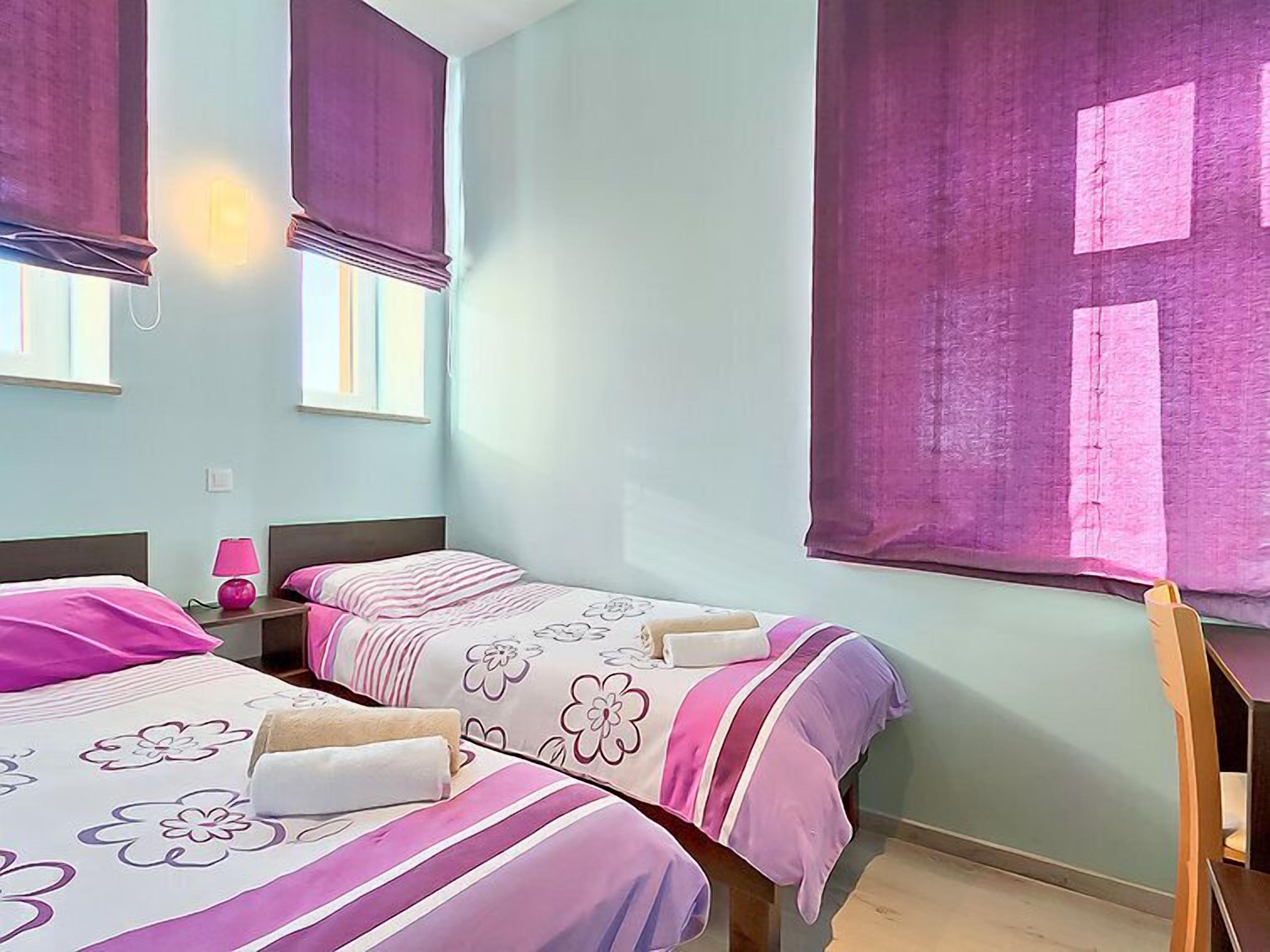 A room with two beds, each with floral-patterned bedding, a nightstand with a lamp, and a large window with purple curtains.