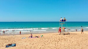 Una playa con socorristas en una torre, gente nadando y caminando sobre la arena, y un cielo azul despejado.