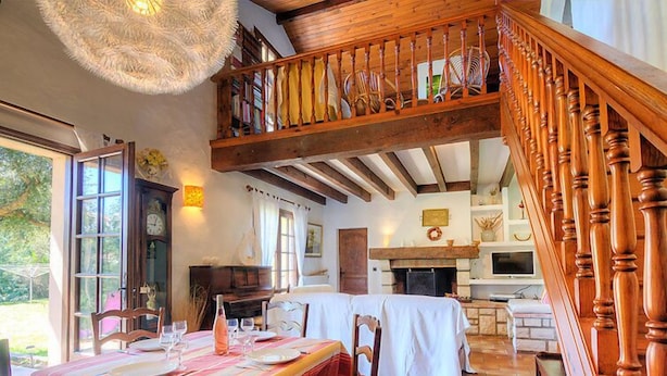 Un comedor con techo de madera alto y una escalera. Hay una mesa preparada con platos y vasos, y un piano al fondo.