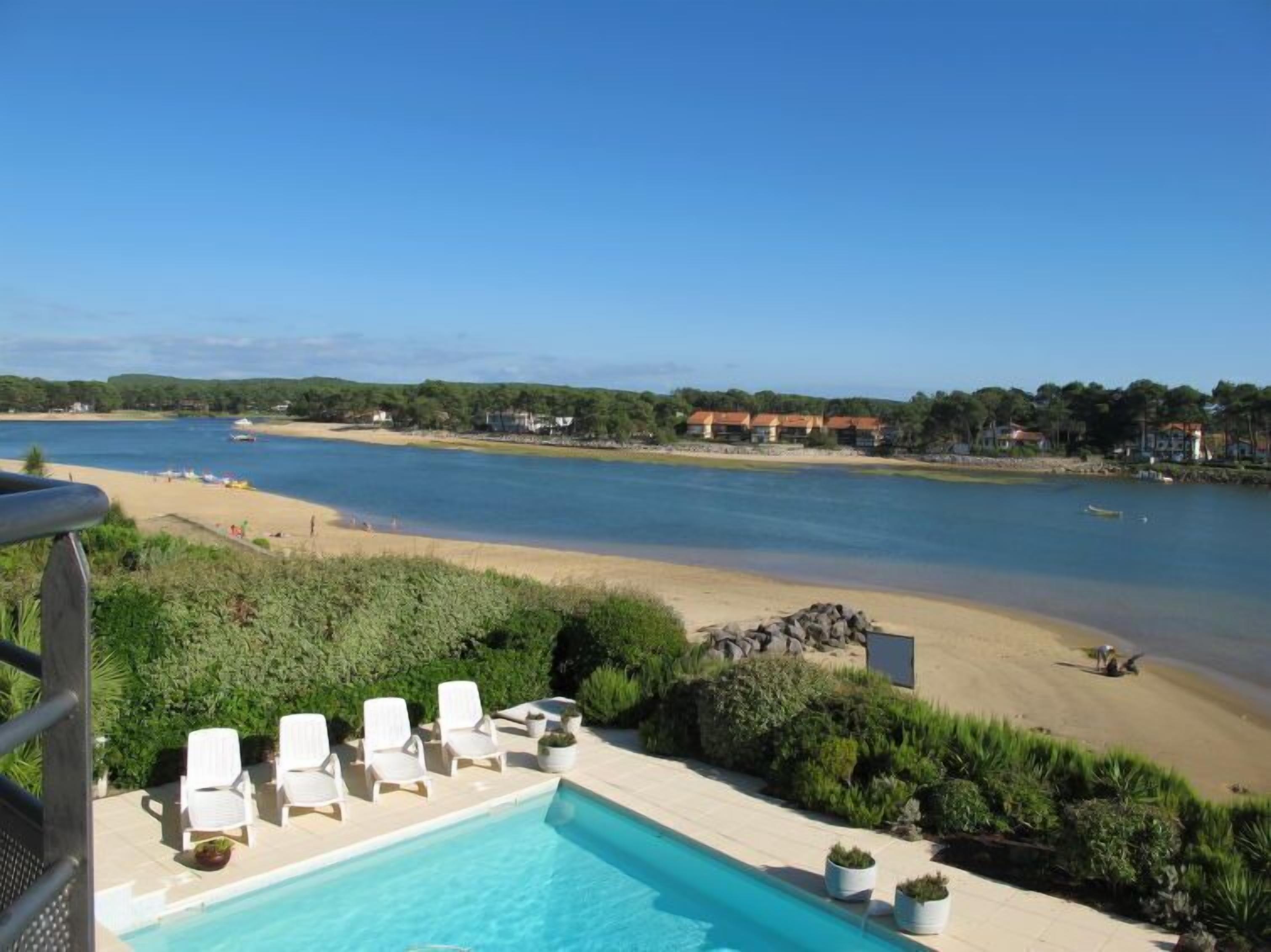A pool area with lounge chairs and a view of a beach and water body.