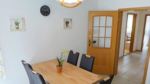 Uma área de refeições com uma mesa de madeira e cadeiras pretas, um relógio e duas plantas em vasos na parede.