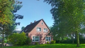 Uma casa de dois andares de tijolo com telhado inclinado, cercada por um gramado bem cuidado e árvores.
