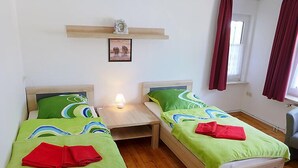 Quarto com duas camas de solteiro, mesa de cabeceira com abajur, prateleira com relógio e quadro, e janela com cortinas.