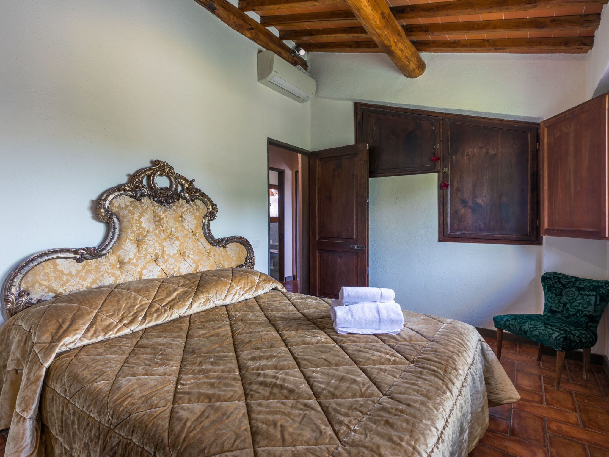 Une chambre avec un grand lit, des poutres apparentes en bois au plafond, une armoire et un fauteuil vert.