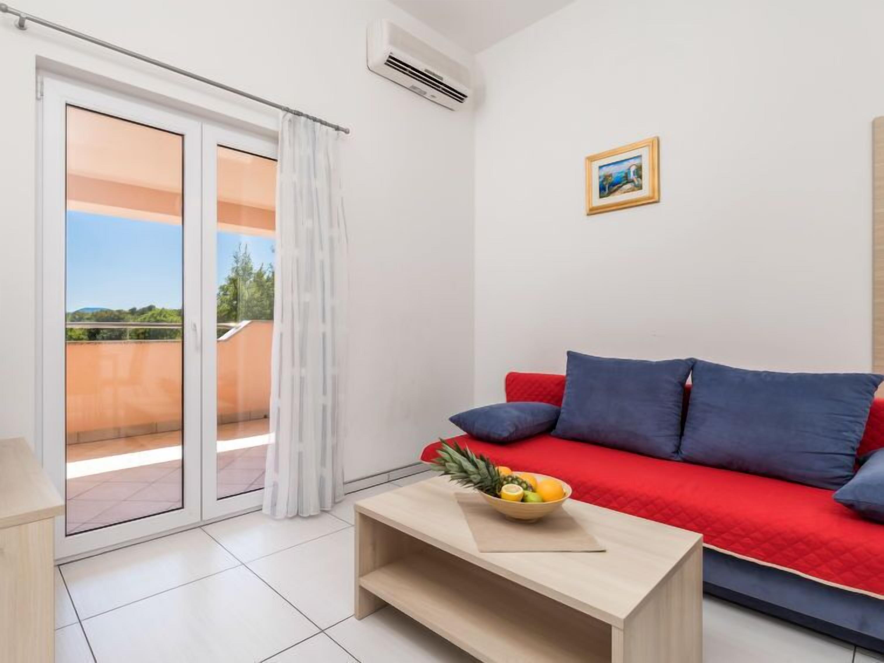 A modern living room with a red sofa, a wooden coffee table with a fruit bowl, and a sliding glass door leading to a balcony.