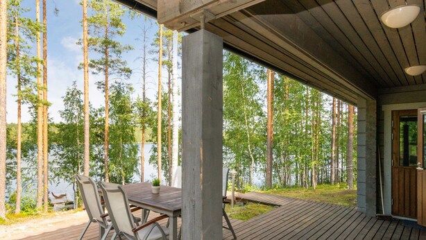 A covered outdoor deck with a table and chairs, overlooking a forest and a body of water. - Hot pool cottage ainu by Interhome (Enonkoski)