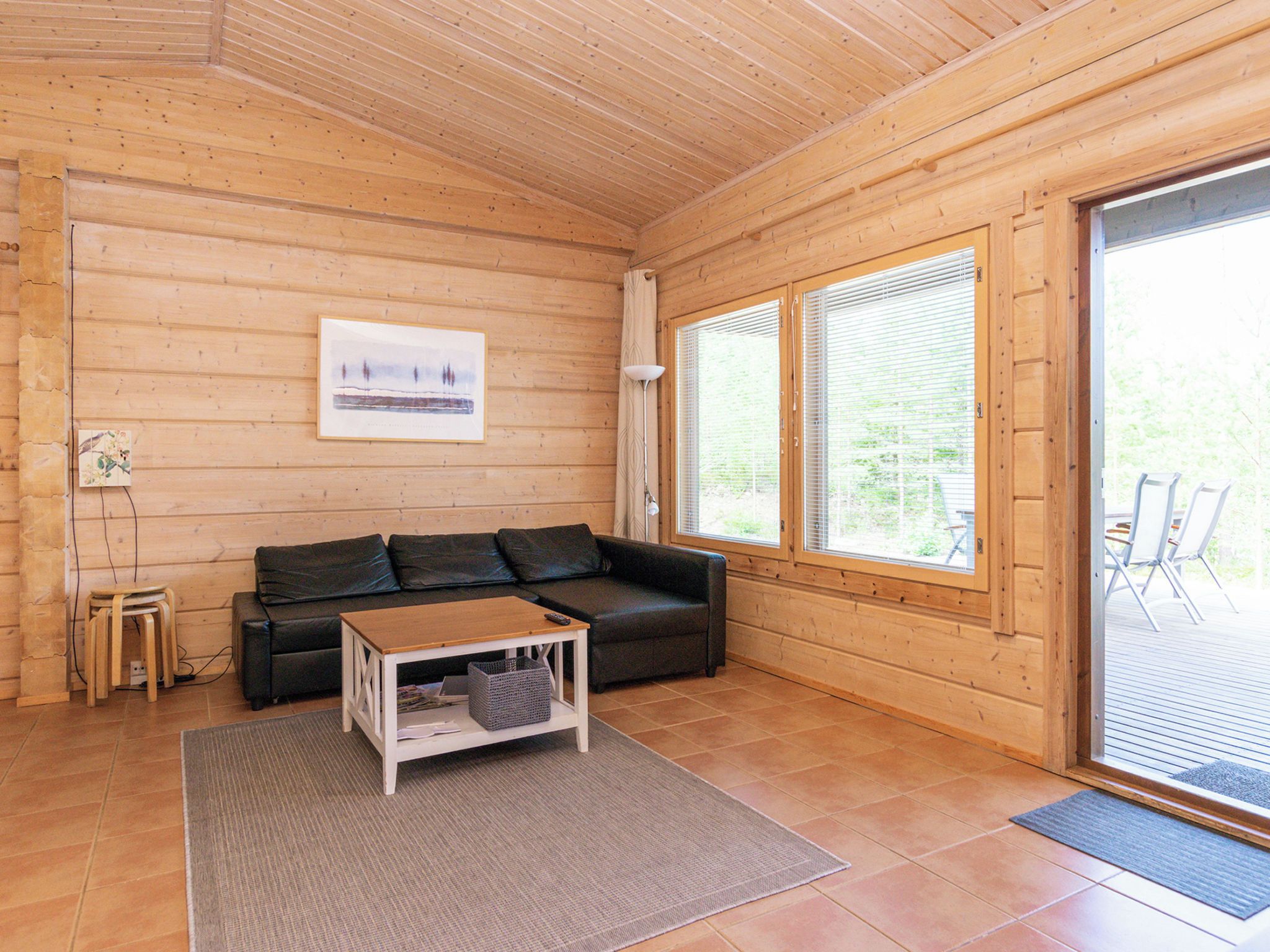 Ein gemütliches Zimmer mit Holzboden, einer schwarzen Couch, einem kleinen Holztisch und einem großen Fenster mit Jalousien.