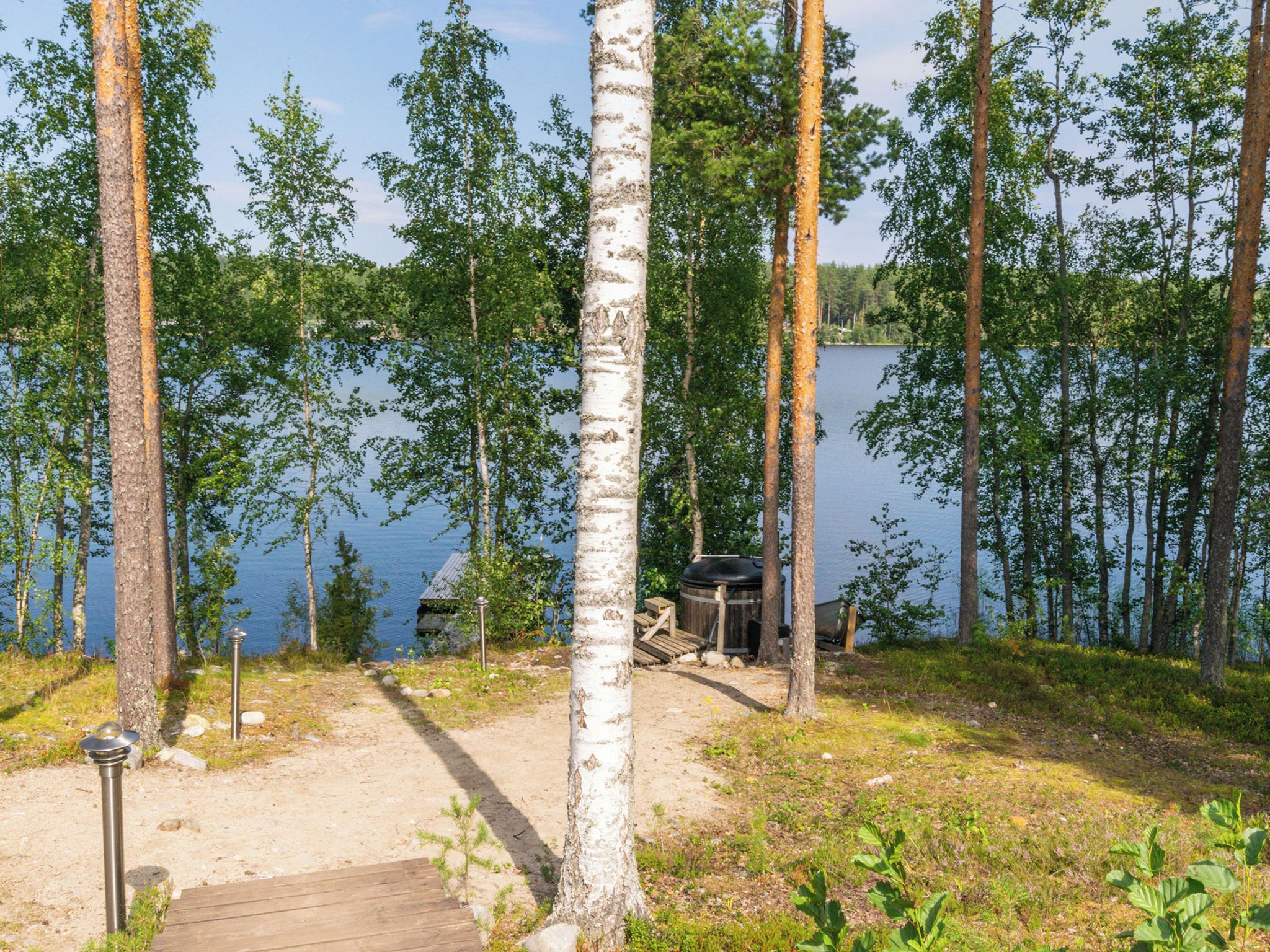 A forested area with a wooden dock leading to a small dock, a trash bin, and a water body in the background.