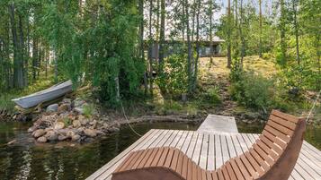 Une terrasse en bois avec un banc incurvé donnant sur une rivière paisible, entourée d’une forêt dense et, au loin, une petite maison.