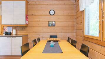 A wooden cabin interior with a dining table, chairs, a clock, and a window with blinds.