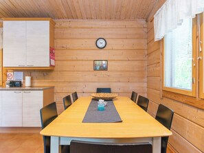 A wooden cabin interior with a dining table, chairs, a clock, and a window with blinds.