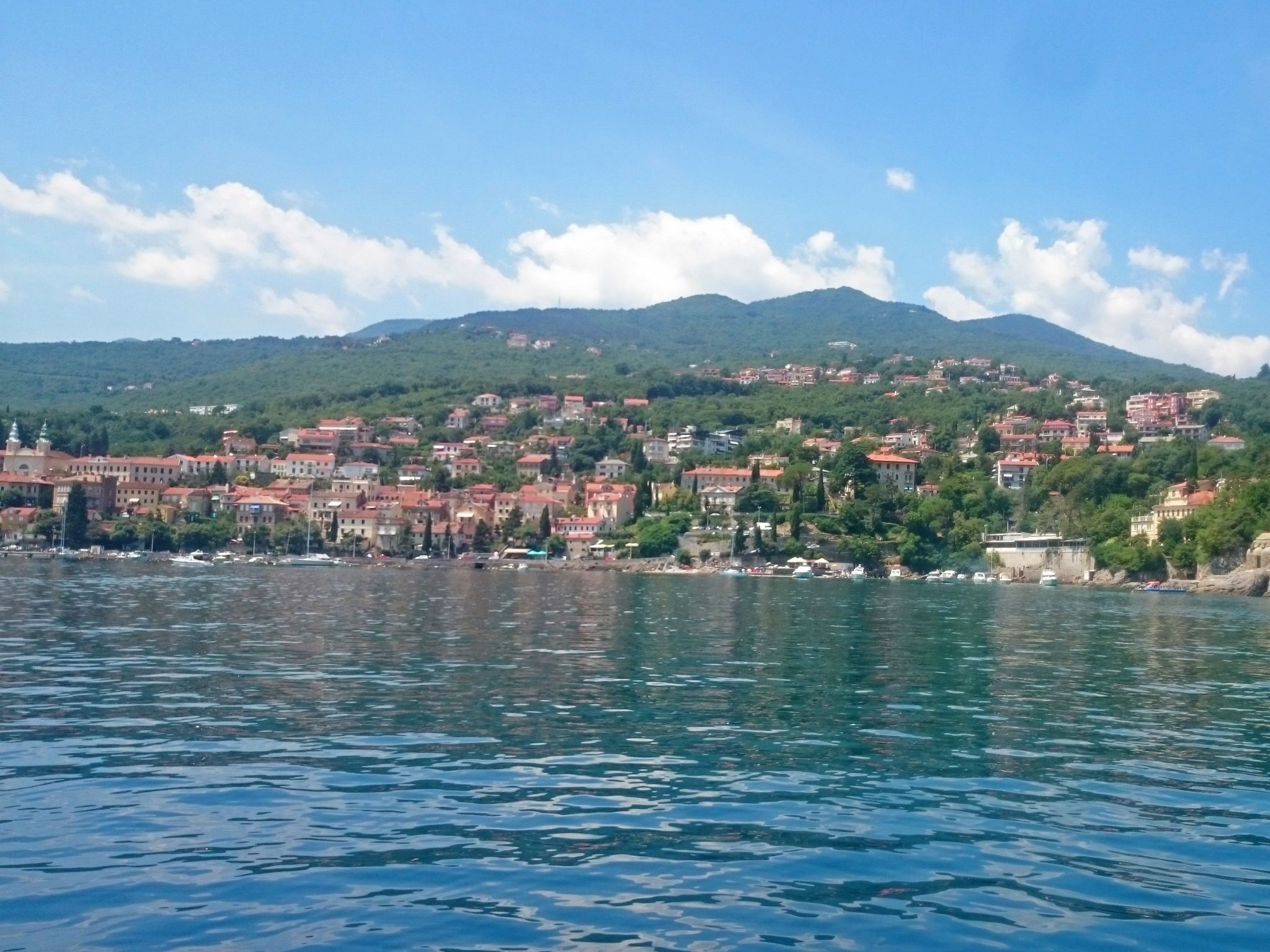 A coastal town with buildings along the shoreline, a body of water in the foreground, and hills in the background.