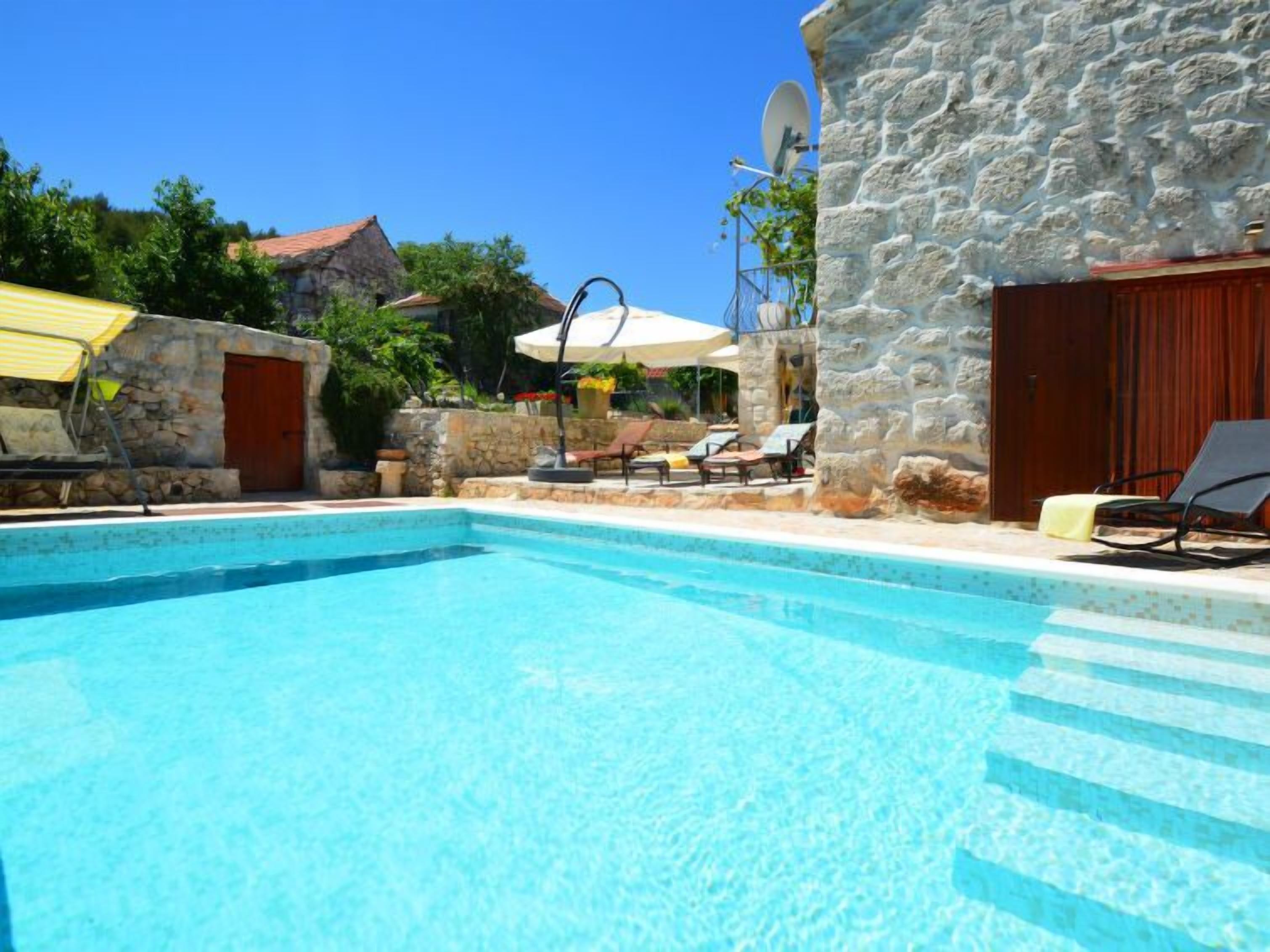 Une piscine d’un bleu éclatant, bordée de chaises longues et d’ombrelles, et entourée de murs de pierre et de végétation.