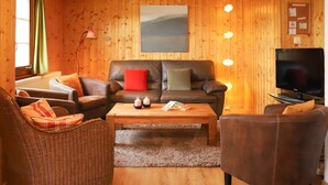Een gezellige woonkamer met een bruine leren bank, een rieten stoel en een bruine fauteuil. Er staat een houten salontafel met boeken en kaarsen. Een flatscreen televisie staat op een standaard aan de rechterkant.