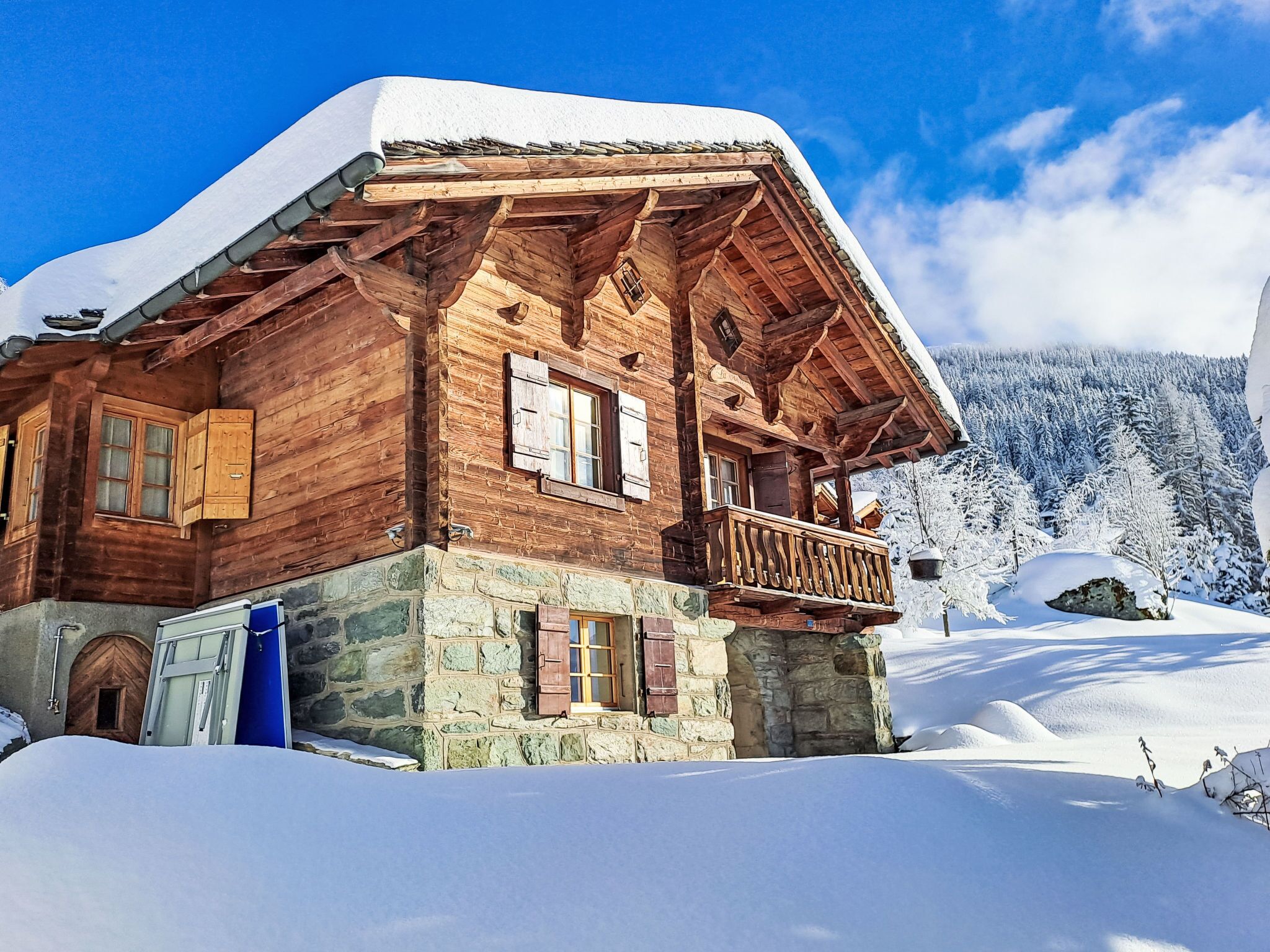 A wooden chalet with a stone foundation, a balcony, and a snow-covered roof.