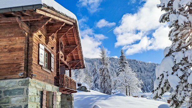 Een houten chalet met een stenen fundering, een balkon en een besneeerd landschap met bomen.