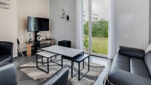 A modern living room with a sofa, a coffee table, and a TV. There is a sliding glass door leading to a balcony with a view of greenery. - La Coloniale by Interhome (Auray)