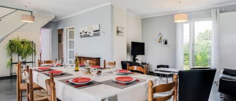A dining area with a long table set for six, surrounded by wooden chairs. A television is mounted on the wall, and a plant is visible in the corner.