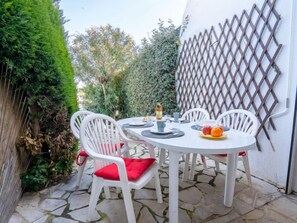 Un pequeño patio al aire libre con una mesa redonda y sillas blancas, rodeado de follaje y un muro ajardinado.
