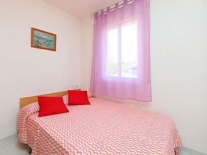 A bedroom with a bed, two red pillows, a window with purple curtains, and a painting on the wall. - Vacation home Mar by Interhome (L'Ampolla)