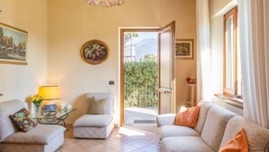 Una sala de estar con un sofá, un sillón y una mesa de centro de vidrio. Hay un cuadro en la pared y una puerta que da al exterior.